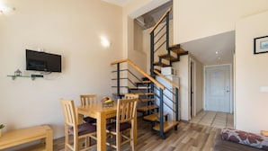 A dining area with a wooden table and chairs, a TV mounted on the wall, and a staircase with wooden steps and black railings.
