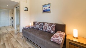 A living room with a brown sofa, patterned cushions, a framed picture on the wall, and a bedside table with a lamp.