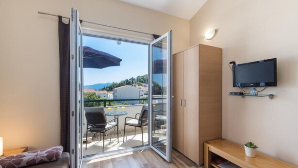 A modern room with a sliding glass door leading to an outdoor balcony, a TV mounted on the wall, and a wooden bench. - Apartments Nmn - Studio Apartment with Balcony 3 (Drvenik)