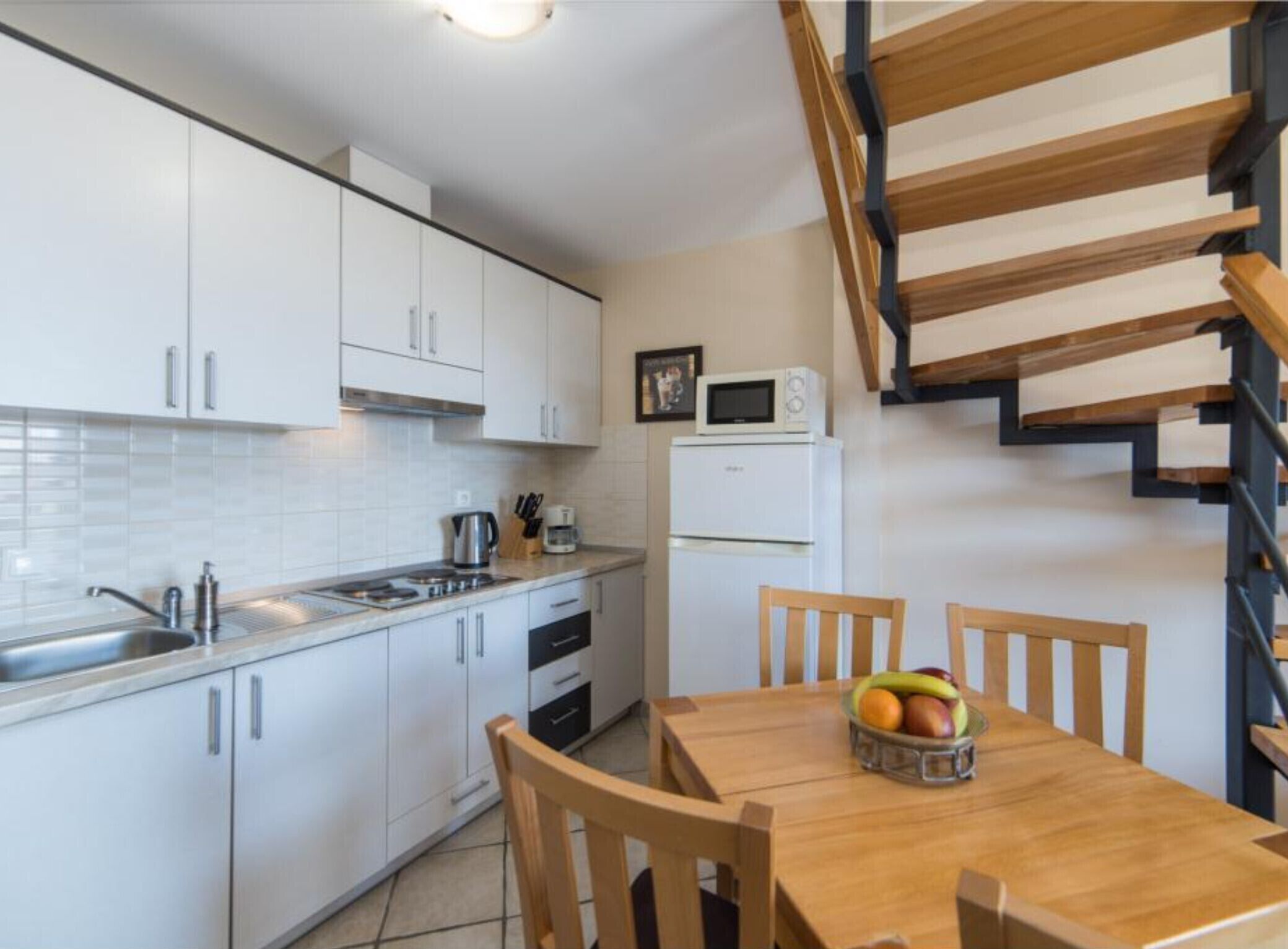 A modern kitchen with white cabinets, a wooden dining table with chairs, a fruit bowl, and a staircase with wooden steps.