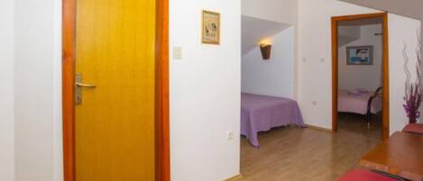 A room with a wooden door, a dining table with chairs, a bed with a purple bedspread, and a small table with a lamp.