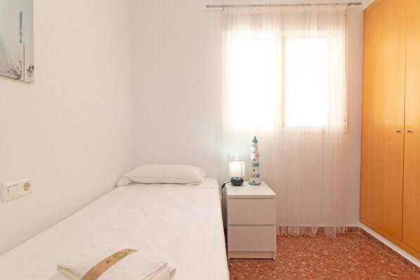 A bedroom with a bed, a nightstand, a lamp, a wardrobe, and a window with sheer curtains.