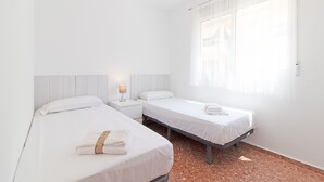 A hotel room with two beds, a nightstand, a lamp, and a window with sheer curtains.