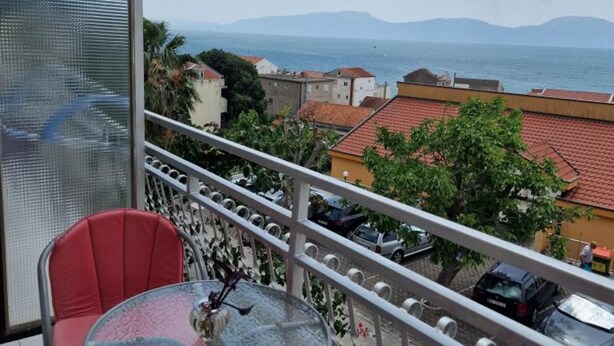 A balcony with a glass table and red chair, overlooking a coastal town with red-roofed buildings and parked cars.