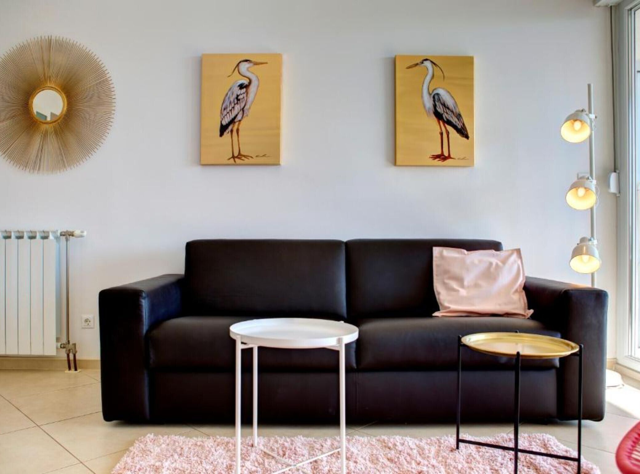 Ein modernes Wohnzimmer mit einem schwarzen Sofa, zwei Vogelgemälden, einem runden Spiegel, einem kleinen weißen Tisch und einer Stehlampe.