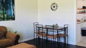 A dining area with a table and chairs, a ceiling fan, a clock, and a painting of a forest on the wall.