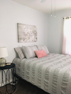 A bedroom with a bed, pillows, a bedside table, a lamp, and a wall-mounted artwork.