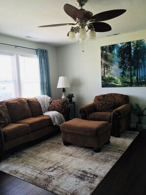 A living room with a brown sofa, a brown armchair, a ceiling fan, a lamp, and a forest-themed wall painting. - Park View Cottage Near Carey Park (Hutchinson)
