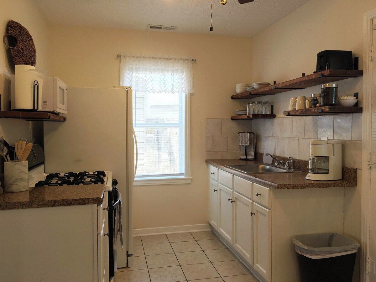 A kitchen with a stove, microwave, refrigerator, and a window with a curtain. There are shelves above the counter holding various kitchen items.