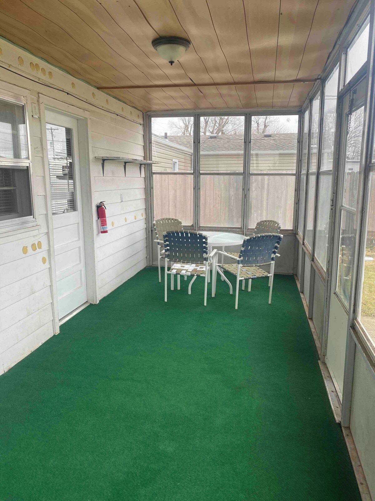 Un patio couvert avec un sol recouvert d’une moquette synthétique verte, deux chaises en métal, une petite table ronde et un extincteur fixé au mur.