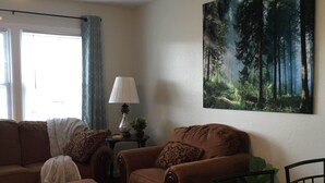 Una sala de estar con sofá, sillón, reposapiés y una mesa de comedor con sillas. Hay un ventilador de techo, una lámpara y un cuadro de un bosque en la pared.
