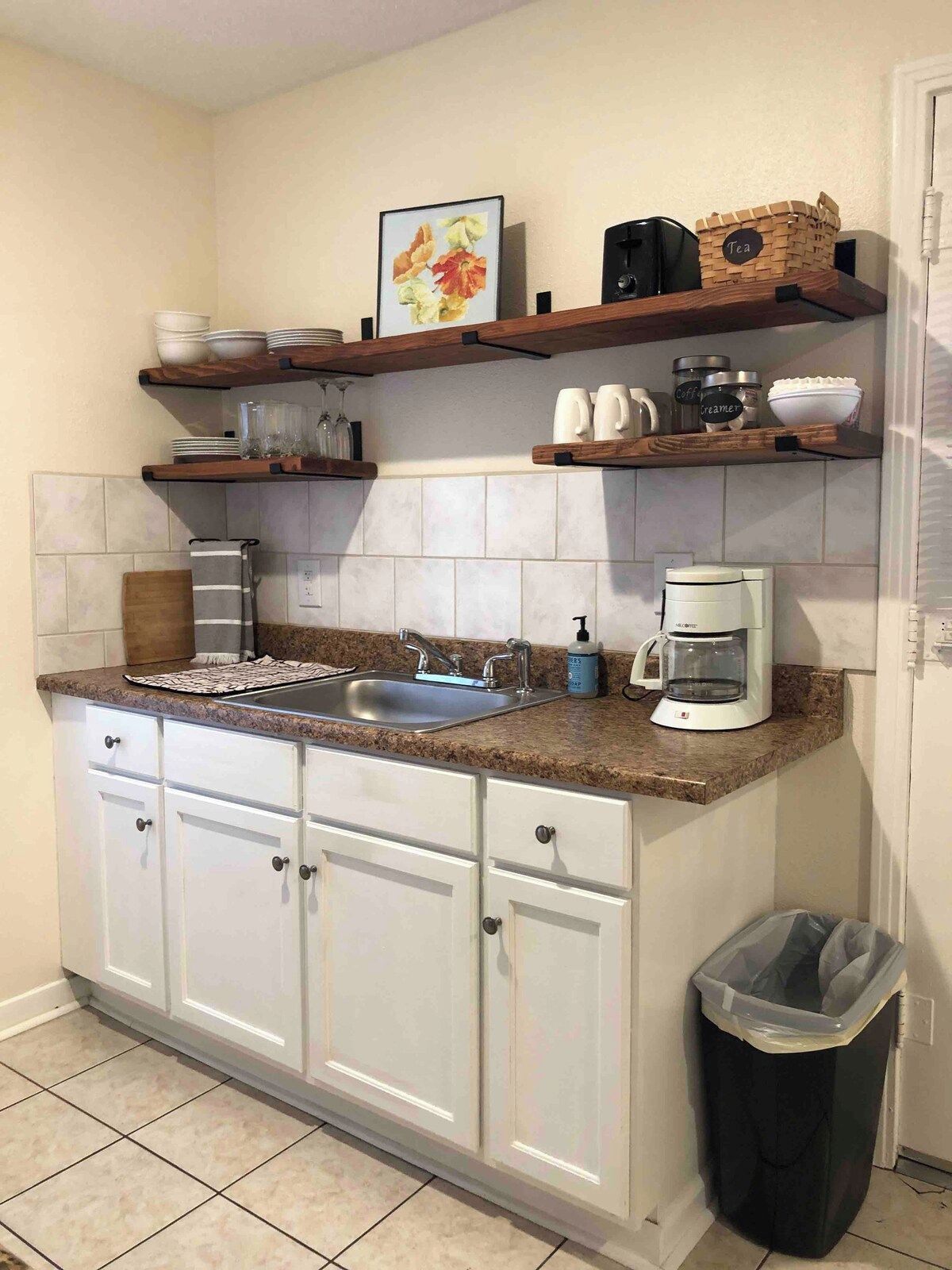 Una cocina con encimera de granito, armarios blancos, cafetera y cubo de basura.