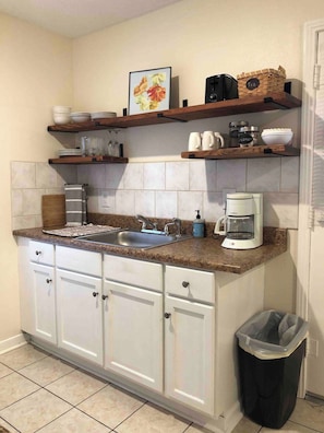 Una cocina con encimera de granito, armarios blancos, cafetera y cubo de basura.