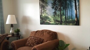 Una sala de estar con un sillón marrón, una mesita con una lámpara, un cuadro grande con motivos de bosque en la pared y una planta en maceta.