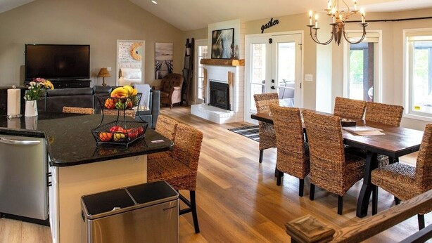 A spacious living area with a fireplace, dining table with wicker chairs, and a kitchen area with a TV and fruit basket. - River front paradise,  boat, kayak, swim,  go to the racetrack, have fun, relax! (Cayuga)