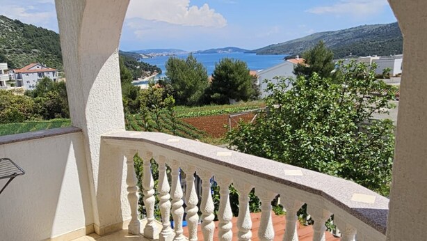 A balcony with a stone balustrade overlooking a coastal landscape with houses and greenery.