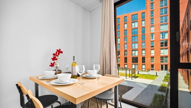 Un comedor moderno con una mesa de madera dispuesta para dos personas, una botella de vino y un jarrón con flores rojas.