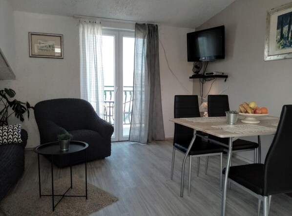 Una sala moderna con sofá, una pequeña mesa de centro, una mesa de comedor con sillas, televisor montado en la pared y un balcón con vistas.