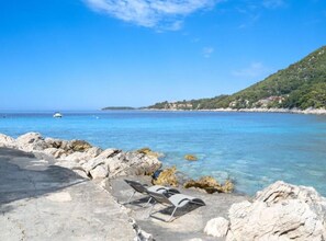 A rocky beach with clear blue water, a few lounge chairs, and a distant view of a hilly coastline with buildings. - Holiday Home Seafront Oleandar (Blato)