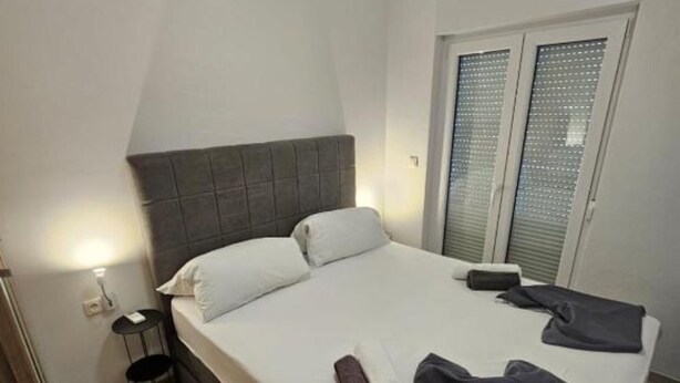 Ein ordentlich bezogenes Bett mit Kissen und Decke, ein kleiner Nachttisch mit Lampe und ein Fenster mit Jalousien.