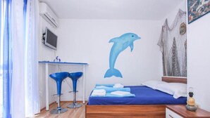 Une chambre avec un lit, un bureau, deux tabourets de bar bleus, une fresque murale représentant un dauphin et une fenêtre avec des rideaux.