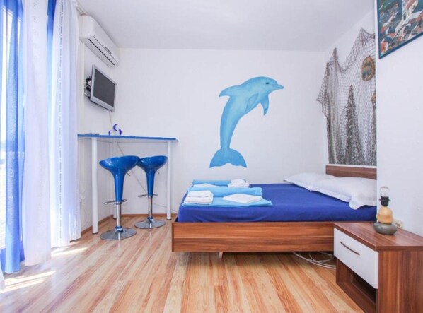 A bedroom with a bed, a desk, two blue bar stools, a wall mural of a dolphin, and a window with curtains.