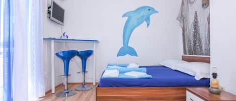 A bedroom with a bed, a desk, two blue bar stools, a wall mural of a dolphin, and a window with curtains.