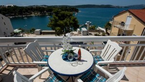 A balcony with a round table, white chairs with plaid cushions, and a view of a coastal town and sea. - Apartments Tomi - Studio With Sea View (2) (Trogir)