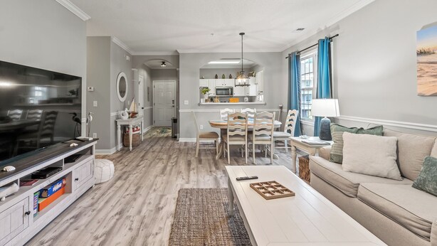 A modern living room with a flat-screen TV, a dining area with a table and chairs, and a kitchen in the background.
