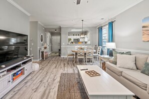 A modern living room with a flat-screen TV, a dining area with a table and chairs, and a kitchen in the background. - Beautifully  Decorated 2nd  floor 2 bedroom Arbor Trace (North Myrtle Beach)
