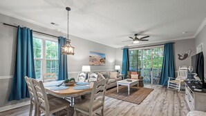 A spacious living room with a dining area, large windows with blue curtains, a ceiling fan, and a view of greenery outside.