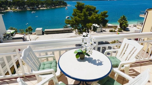 A balcony with a round table and white chairs, overlooking a coastal area with boats and a clear view of the sea. - Apartments Tomi - Double Room With Sea View (3) (Trogir)