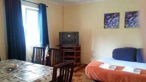 A room with a dining table, chairs, a television on a stand, a bed with a blue blanket, and two floral paintings on the wall. - Apartments Štrbić - One Bedroom - Nina (Karin Gornji)