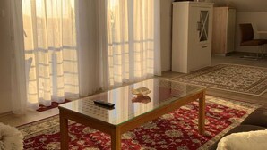 A living room with a glass-top coffee table, red patterned carpet, white curtains, and a white cabinet. - Apartment Bordoa de Macura with Bay View (Zadar)