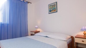 A bedroom with a bed, two bedside tables, a nightstand, a window with blue curtains, and a framed picture on the wall.