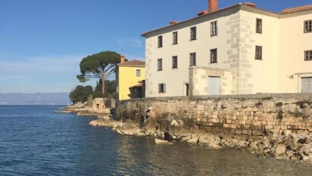 A coastal building with a stone facade, a yellow building adjacent to it, and a clear view of the sea.