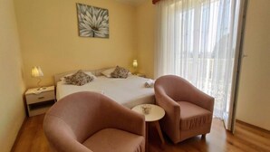 Una habitación de hotel con una cama, dos sillones, una mesita, una mesita de noche con lámpara y una ventana con cortinas translúcidas.