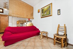 Une pièce avec un canapé rouge, une chaise en bois, un petit tabouret, un meuble en bois, une lampe en osier et un tableau encadré accroché au mur.