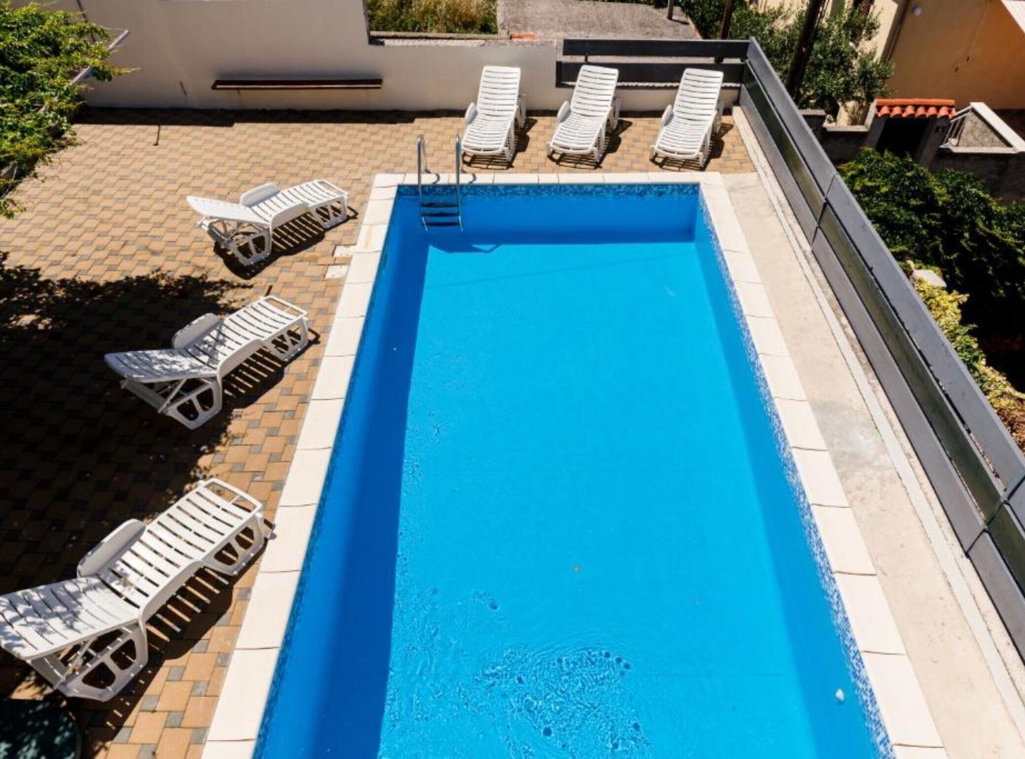 A rectangular swimming pool with a tiled edge, surrounded by white lounge chairs and a paved deck.