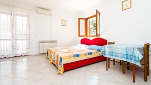 Une chambre avec un lit rouge, une table à carreaux bleus et blancs, un radiateur et une fenêtre avec des rideaux légers.