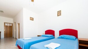 Ein Hotelzimmer mit zwei Betten, jeweils mit rotem Kopfteil und blauer Bettwäsche. An der Wand befinden sich zwei gerahmte Bilder, eine Holztür und ein gefliester Boden.