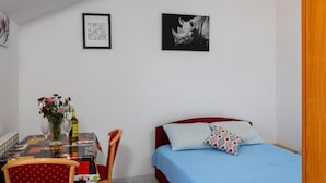Une petite pièce bien éclairée, avec un lit, une table entourée de chaises et deux tableaux encadrés accrochés au mur.