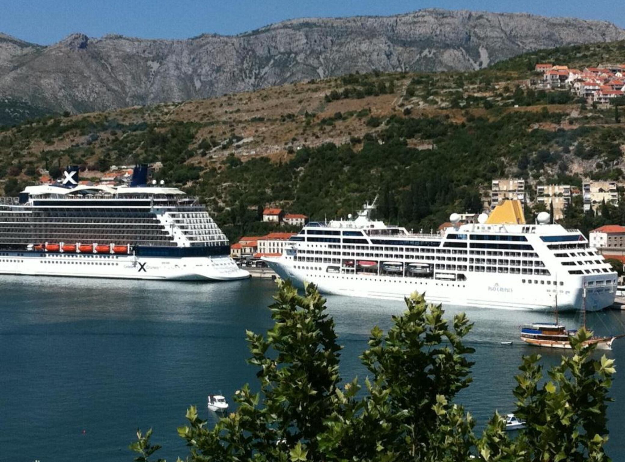 Dos grandes cruceros amarrados en un puerto con montañas al fondo y un pueblo pequeño a lo lejos.