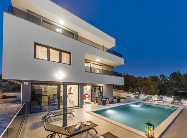 Una casa moderna de dos pisos con piscina, mobiliario exterior y una entrada bien iluminada.