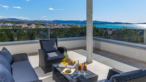 Een balkon met rieten meubilair, een tafel met drankjes en fruit, en uitzicht op een kustplaatsje en zee.