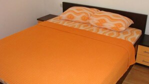Una cama con una manta naranja y almohadas estampadas, una mesita de noche de madera y un mueble con cajones.