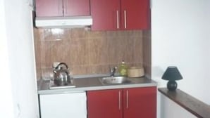 Een kleine keuken met rode kastjes, een witte koelkast, een waterkoker, een wastafel en een lamp op een houten plank.