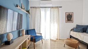 A modern living room with a blue accent wall, a large flat-screen TV, a blue armchair, a wicker ottoman, and a bed with blue pillows.