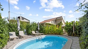 A swimming pool with a tiled edge, surrounded by lounge chairs and umbrellas, with a house and vegetation in the background.