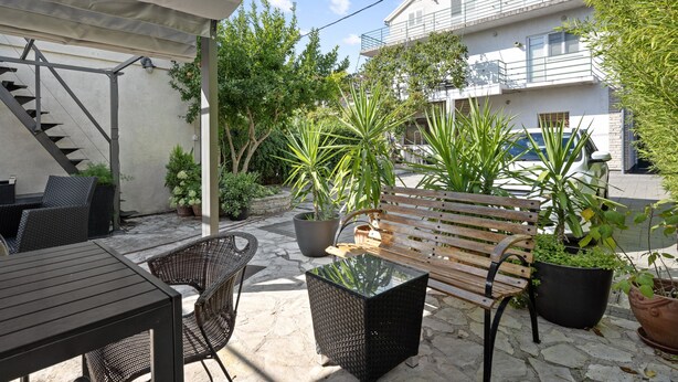 An outdoor patio area with a wooden bench, wicker chairs, and a glass table. There are potted plants and a staircase leading to an upper level.